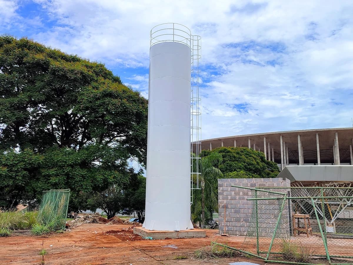 Reservatório metálico tubular vertical na cor branca com escada de acesso lateral e guarda-corpo superior sobre base de concreto